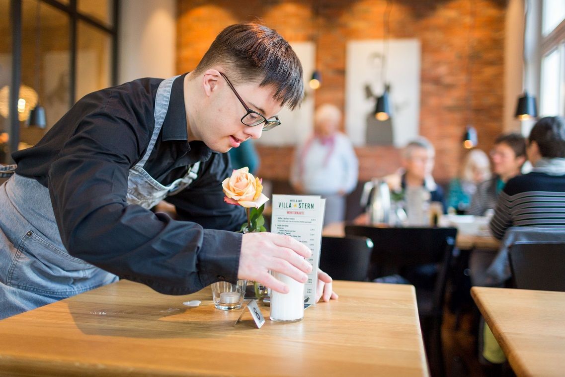 Junger Mann mit Downsyndrom deckt in Restaurant einen Tisch ein (nachhaltige Unternehmen beschäftigen Menschen mit Behinderung)