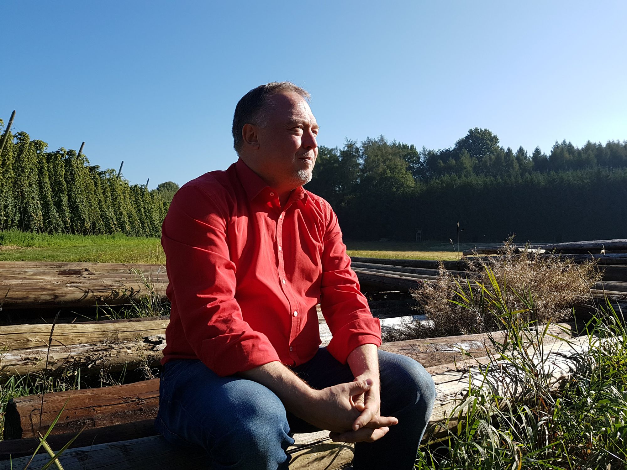 Mann Mitte 50, rotes Hemd und Jeans, sitz auf einem Baumstamm in schöner Naturlandschaft unter blauem Himmel