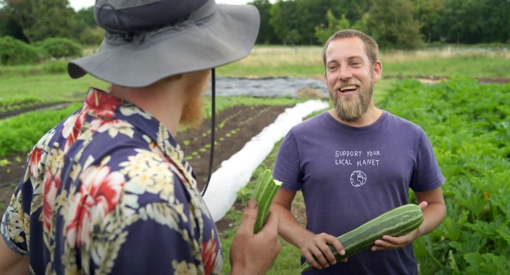 Junger Bauer mit blondem Kurzhaar und Vollbart hält eine große Zucchini in der Hand und lacht